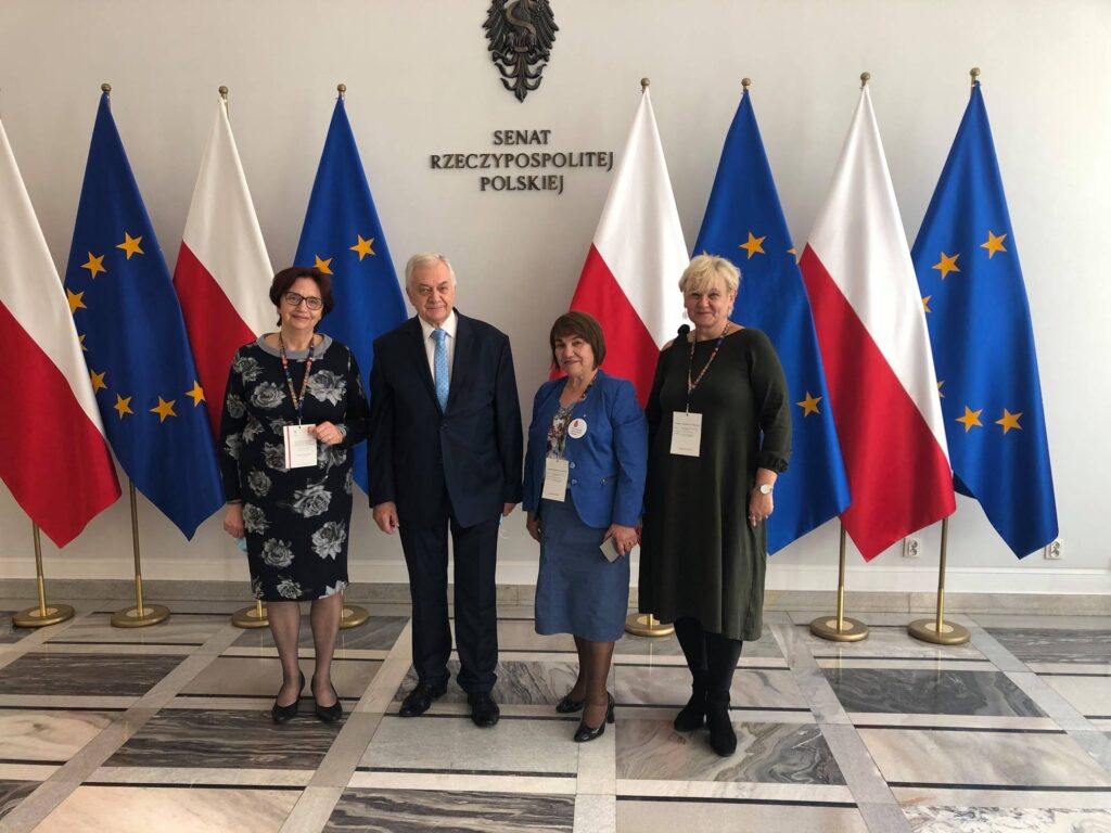 Trzy kobiety i mężczyzna w siedzibie senatu. Pozują na tle flag Polski I Unii