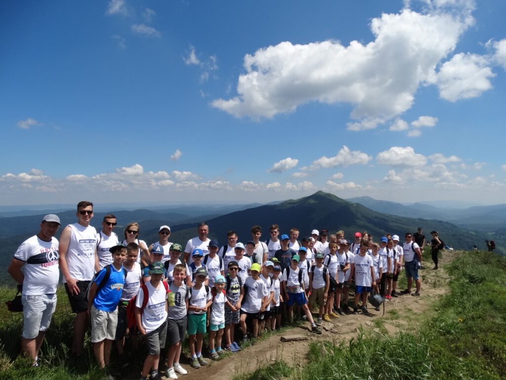 Duża grupa dzieci i młodziezy w białych bluzkach i krótkich spodnaich stoi na ścieżce. W tle szczyty gór.