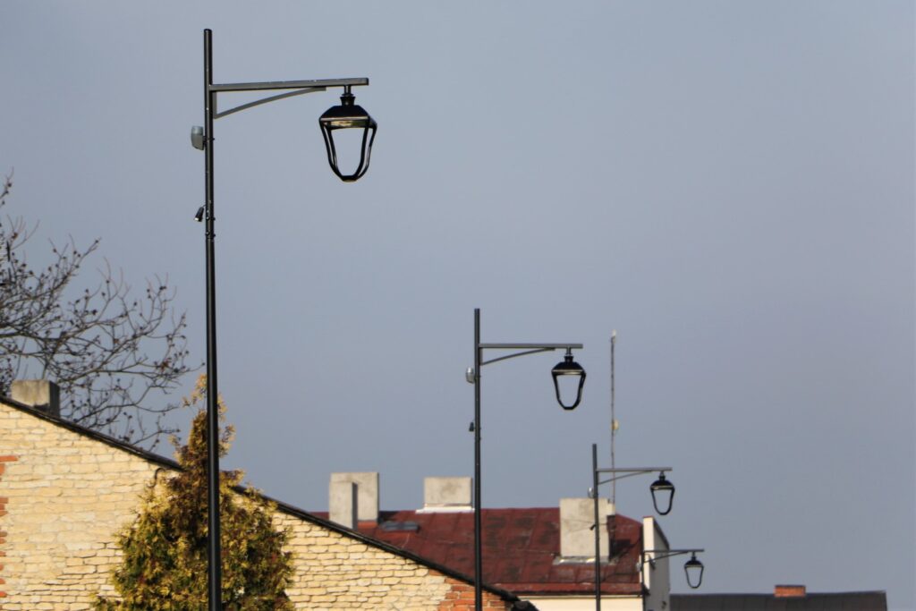 Rząd nowoczesnych latarni ulicznych. Slupy i oprawy w kolorze czarnym. W tle dachy domów