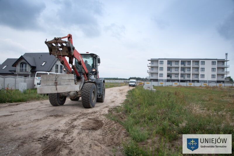 Maszyna budowlana jedzie po polnej drodze. Na drugim planie blok mieszkalny i biały samochód